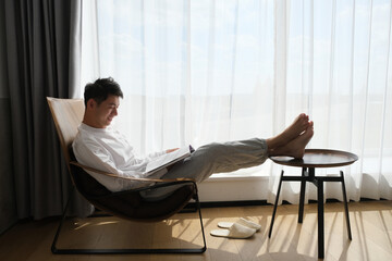 smiling Asian young man reading book on chair near window under sunshine