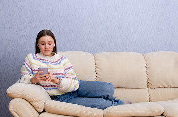 Young teen girl resting on a couch, using smartphone digital device at home. Texting, social media, online shopping.