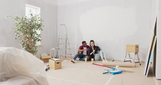 Handsome man sits on the floor with his beautiful sweetheart, taking a break from painting apartment walls during a renovation, drinking take-out coffee, browsing inspiration on a tablet