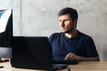 Handsome serious bearded man sitting at the office desk, working on laptop, looking at computer screen against concrete wall in modern office