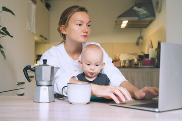 Young woman studying or working online at home while having breakfast with her baby on kitchen. Millennial mother on maternity leave with child. Freelancer busy mom with laptop searching information.