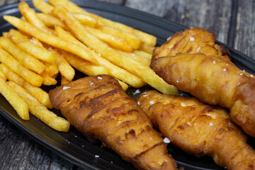 Battered chicken fillets with french fries, seasoned with sea salt, on a black stoneware plate.  On a wooden background