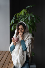Sick woman cough suffer from seasonal flu, cold or coronavirus disease drink hot tea shivering stand at radiator near window. Ill young female with sickness at home. Healthcare and medication concept