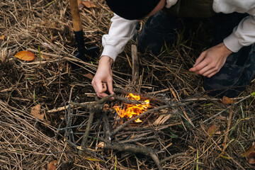 Close-up high-angle view of unrecognizable survivalist male putting kindling on burning fire to keep warm and cook food at outdoors on overcast cold day. Concept hiking and traveling outdoor.