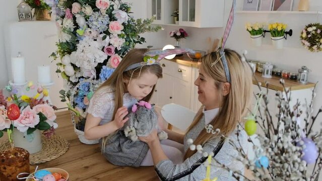 A cute little girl in bunny ears with her mom is holding a rabbit while sitting at a table in the kitchen. Decorations for the celebration of Easter