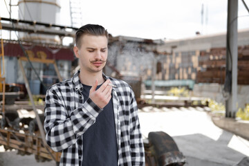 young handsome man with mustache in checkered black and white shirt smoking a cigarette outside....