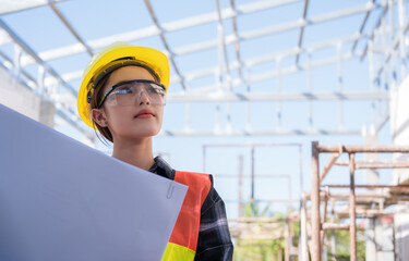 Asian woman civil engineer wears safety yellow helmet with goggles and holding blueprint while...