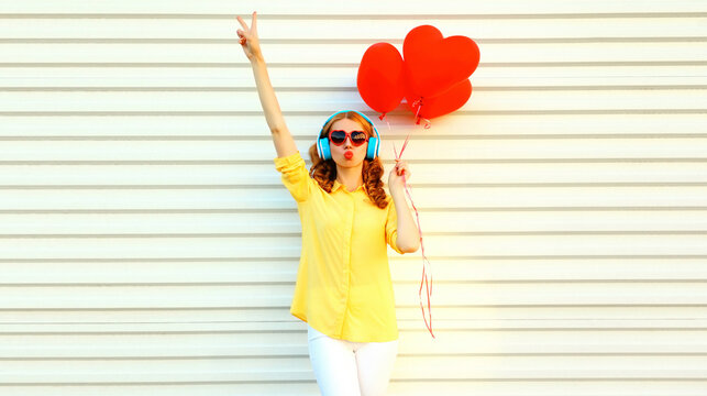 Portrait of young woman with bunch of red heart shaped balloons listening to music in headphones and blowing her lips sending sweet air kiss on white background
