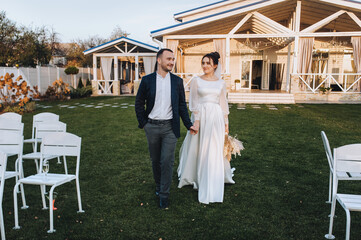 Fototapeta na wymiar A stylish groom and a beautiful brunette bride in a white dress with a bouquet of reeds are walking, holding hands, along the green grass against the backdrop of chairs, a house, a cottage.