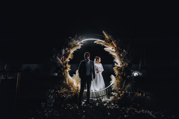 A stylish groom and a beautiful bride in a long dress are standing at night near a reed arch...