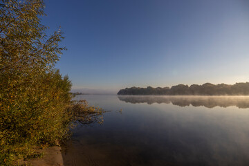 Autumn landscape in the early morning on the river. Misty water surface. Yellow leaves on trees and bushes are illuminated by the rays of the rising sun. Dawn on a cold autumn morning.