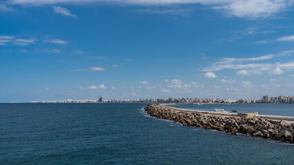 Seascape in Alexandria. A stone pier in the Mediterranean Sea, yachts are visible. In the distance, on the horizon, there is an embankment with city buildings. Blue sky with clouds. Egypt
