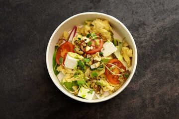 Bowl with scramble, feta cheese, tomatoes, walnuts and microgreens on a dark background in an eco paper box. Healthy food delivery concept.