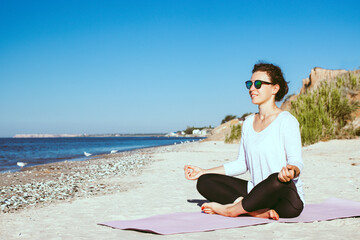Naklejka na ściany i meble Yoga on the beach. Smiling young woman sitting in lotus pose, practicing breathing techniques near ocean on blue sky background. Meditation and pranayama. Summer healthy lifestyle. Copy space.