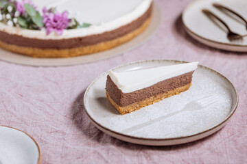 Chocolate cheesecake on bright plate with whole cake in blurry background. Decorated with flowers on pastel purple cloth. Vintage atmosphere, festive also. Juicy cake, one piece