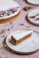 Chocolate cheesecake on bright plate with whole cake in blurry background. Decorated with flowers on pastel purple cloth. Vintage atmosphere, festive also. Juicy cake, one piece