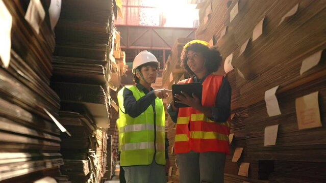 Engineer asian and african woman wearing safety helmet and vest holding clipboard and take note in the automotive part warehouse.Products and corrugated cardboard. Factory for the manufacture.