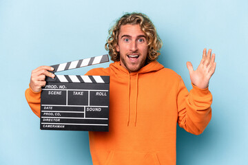 Young caucasian man holding a clapperboard isolated on blue background receiving a pleasant...