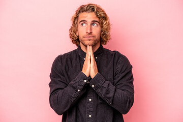 Young caucasian man isolated on pink background praying, showing devotion, religious person looking for divine inspiration.