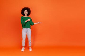 Full length of friendly smiling woman with Afro hairstyle wearing green casual style sweater presenting copy space for advertisement or promotion. Indoor studio shot isolated on orange background.