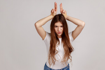 Fototapeta premium Portrait of aggressive bully woman with dark hair showing bull horns gesture over head, frowning as before attack, wearing white T-shirt. Indoor studio shot isolated on gray background.