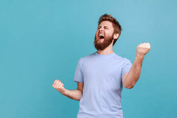 Portrait of overjoyed bearded man standing with excited expression, raising fists, screaming,...