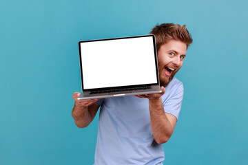 Portrait of bearded man holding laptop with blank white screen and looking at camera with excited...