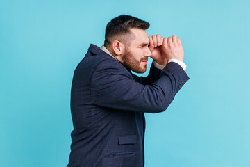 Side view portrait of bearded man wearing official style suit making glasses shape, looking through binoculars gesture with surprised expression. Indoor studio shot isolated on blue background.