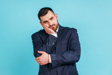 Portrait of sad upset or bored bearded young adult man wearing official style suit standing and looking at camera with dissatisfied sadness face. Indoor studio shot isolated on blue background.