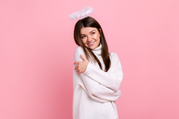Charming young adult brunette angelic woman with nimb over head, looking away and hugging herself, wearing white casual style sweater. Indoor studio shot isolated on pink background.
