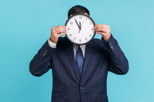Unknown male person wearing official style suit hiding face behind wall clock display, wasting his time, procrastination, organization of working time. Indoor studio shot isolated on blue background.