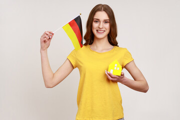 Positive smiling teenager girl in yellow casual T-shirt showing small paper house and holding flag of germany, satisfied with accommodation purchase. Indoor studio shot isolated on gray background.