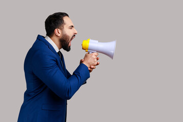 Side view of young adult handsome bearded man screaming in megaphone, announcing important information, wearing official style suit. Indoor studio shot isolated on gray background.