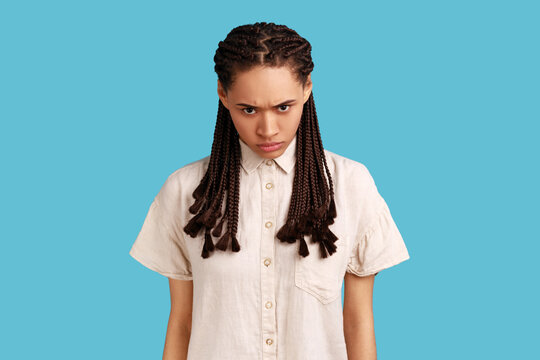 Portrait of aggressive woman with black dreadlocks, having irritation squints face, being annoyed with something, wearing white shirt. Indoor studio shot isolated on blue background.