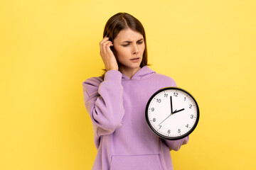 Portrait of confused woman holding in hands big wallclock, has no time , worried about deadline, looking away, wearing purple hoodie. Indoor studio shot isolated on yellow background.