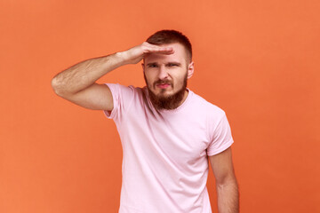 Fototapeta na wymiar Portrait of bearded man looking far away at distance with hand over head, attentively searching for bright future, wearing pink T-shirt. Indoor studio shot isolated on orange background.