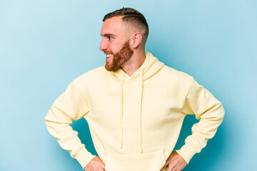 Young caucasian man isolated on blue background confident keeping hands on hips.