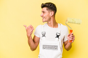 Young painter man isolated on yellow background points with thumb finger away, laughing and carefree.