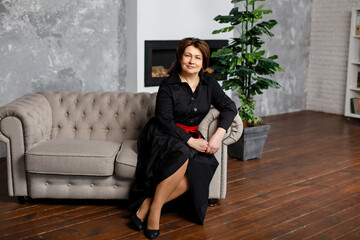 Portrait of busy modern 50 years old middle-aged woman in black dress in an office making plans, looking at camera with positive friendly smile. Female supervisor on workplace. Woman entrepreneur
