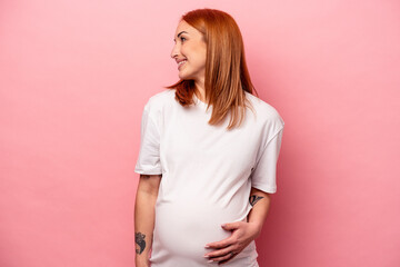 Young caucasian pregnant woman isolated on pink background looks aside smiling, cheerful and pleasant.