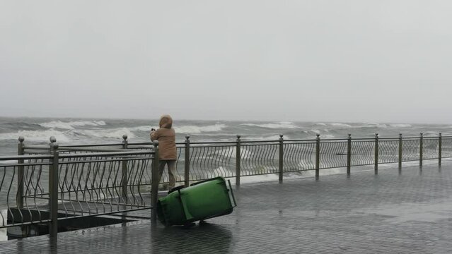 Unrecognizable woman taking pictures of the sea on her phone during a violent storm, she is trying to stay on her feet against the hurricane winds. Storm called Nadia on the Baltic Sea