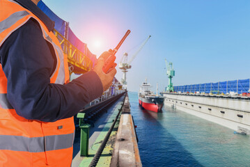 A man holding a walkie talke, or radio, wearing reflective shirt, controls the cargo ship into the dry dock for ship repair, maintenance in shipyard with copy space for texture.