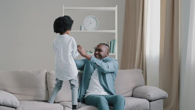 African american father with little daughter spend their free time together having fun playing actively dancing to music caring dad sitting on couch holding kid girl by hands child joyfully jumping