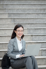 Portrait of happy young businesswoman in glasses sitting outdoors and working on laptop