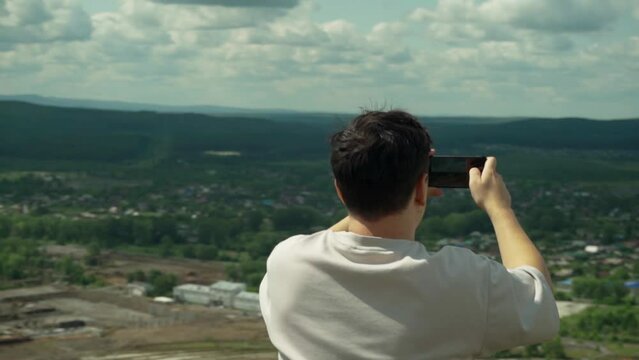 A young adult male takes a smartphone picture from the top of a large hill. 