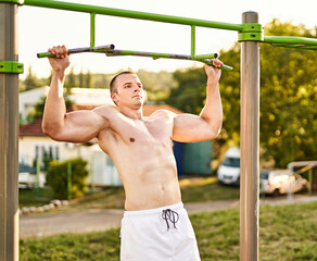 Fototapeta premium Muscular young bodybuilder guy doing pull up exercise on a bar in an outdoor public fitness park - Strength and vitality wellness concept with athletic millennial man - Street workout exercise