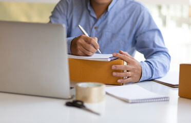 Man writing a notepad of orders and goods from internet laptop.