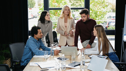 multiethnic business colleagues talking near desk with gadgets and documents in meeting room