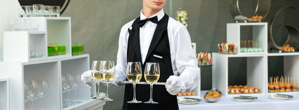 Midsection Of Professional Waiter In Uniform Serving Wine During Buffet Catering Party, Festive Event Or Wedding. Full Glasses Of Champagne On Tray. Outdoor Party Catering Service, Waiter On Duty.