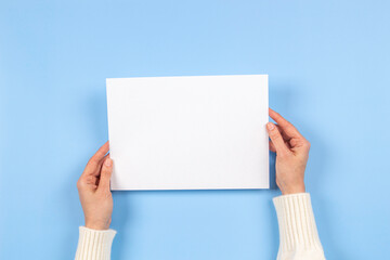 Female hands holding blank white paper sheet on light blue background. Top view. Mockup paper with copy space for text
